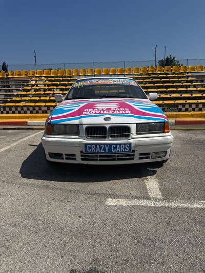 The image depicts a white BMW E36 with custom "CRAZY CARS MOVIEPARK" livery, including a "CRAZY CARS" license plate, parked on asphalt adjacent to yellow stadium seating and a checkered barrier. The location is Općina Funtana, Croatia. No people are visible. The car is casting a shadow on the asphalt. The sky is blue and clear.