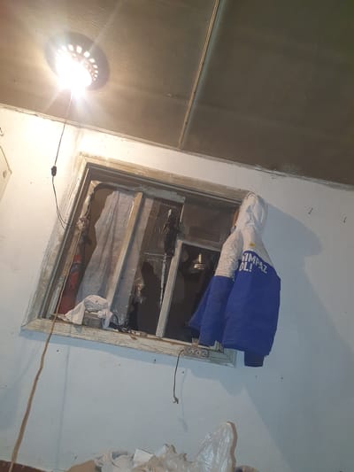 The image depicts a dimly lit interior room with a broken window. A ceiling light fixture illuminates the scene from the upper left. The walls are painted white, and the ceiling appears to be made of paneling. A framed window, with its glass shattered and partially covered by makeshift materials like plastic sheeting and fabric, is central to the image. A blue and white jacket is hanging on the wall to the right of the window. Wires and cords are visible, including one extending from the ceiling light and another near the window. The scene suggests a state of disrepair or neglect, possibly in a residential or abandoned space. The location is identified as Rassvet, Kazakhstan.