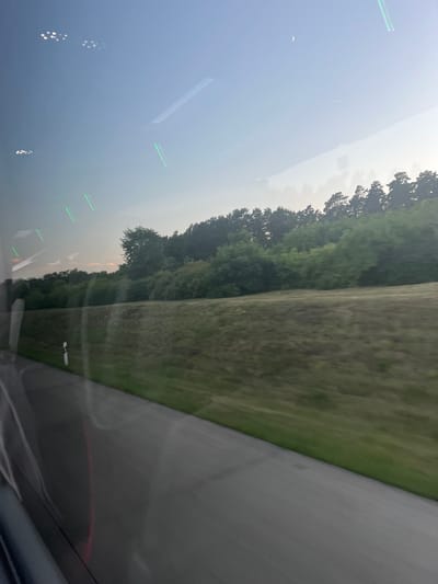 The image captures a view from inside a vehicle, likely a bus or train, looking out a window. The foreground features a blurred asphalt road with a green grassy shoulder. A white and black road marker is visible along the side of the road. Beyond the road, a slope covered in dry grass leads up to a line of lush green trees and dense foliage, suggesting a rural or semi-rural environment. The sky is a pale blue with a sliver of a crescent moon visible in the upper right quadrant. Reflections on the window obscure parts of the scene, showing streaks of light and blurry shapes, including what appear to be distant lights and linear green light sources. The overall impression is one of travel through a landscape during twilight or early morning. The location is identified as Kleinmachnow, Germany.
