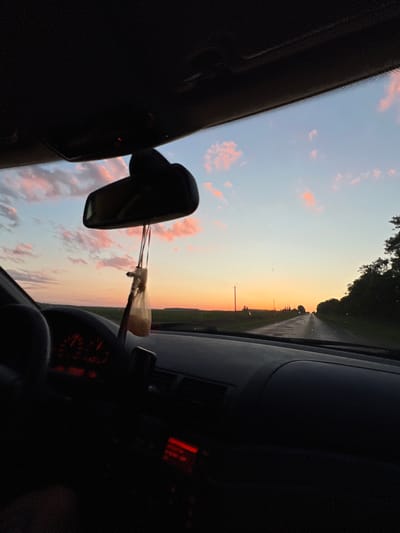 This media file depicts an interior view from a car as it travels along a road in Kypiachka, Ukraine. The scene is dominated by the sky, which displays a gradient of colors from pale blue at the top to a warm orange and pink near the horizon, indicating sunset or sunrise. Scattered pink clouds are visible against the blue sky.
The image is oriented sideways, with the dashboard and windshield on the left, and the right side of the car's interior on the right. A rearview mirror is prominently featured in the center-left of the frame, reflecting the sky. Hanging from the mirror are what appear to be air fresheners.
The road ahead is visible, receding into the distance and bordered by dark green trees on the left and a lighter green field on the right. A utility pole with wires can be seen along the roadside. The car's dashboard is partially visible on the right, with a red illuminated display that is difficult to read. Some of the dashboard instruments with red illumination are also visible.
The overall mood is peaceful and contemplative, capturing the beauty of a rural sunset or sunrise. There is no discernible activity involving people, but the presence of the car suggests travel. The lighting and colors suggest it is either early morning or late evening. No legible text is visible in the image.