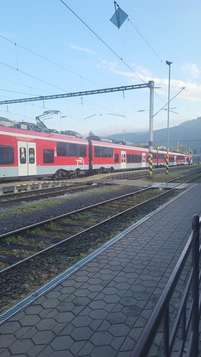 This is a photograph of a red and white train stopped at a train station in Kysak, Slovakia. The train is composed of several cars, and its livery features a white circular logo with a stylized "ÖBB" symbol. The train is on a track that runs parallel to a paved platform. The platform is made of hexagonal grey pavers and has a metal grate running along its edge. Adjacent to the platform are multiple sets of train tracks, with ballast and some vegetation visible between the rails. Overhead, a complex network of electrical wires for the train's power system is suspended from poles. In the background, hills covered in trees are visible under a blue sky with scattered clouds. The lighting suggests it is either early morning or late afternoon. There are no people clearly visible in the frame.