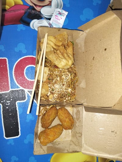 A takeout container holds an assortment of food. The top section features noodles with fried wontons and sprinkled sesame seeds. Chopsticks rest across the top. The bottom section contains three golden-brown fried chicken nuggets. A small packet of sauce sits near the container, along with some cutlery. The meal rests on a blue tablecloth with cartoon character designs.