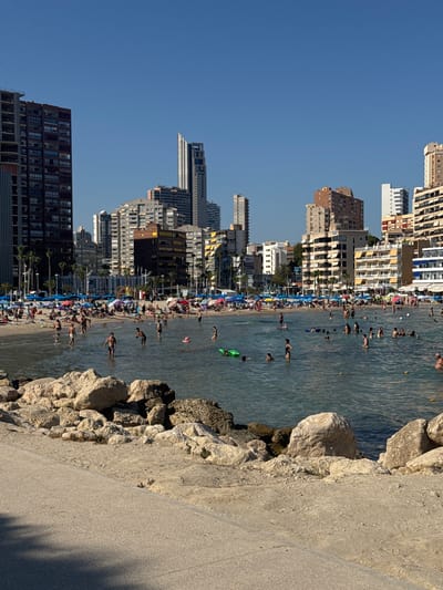 This media file captures a vibrant beach scene with a backdrop of a densely populated, modern city. The image is oriented vertically, with the sky at the top and the water at the bottom. A multitude of people are scattered across the sandy beach and in the shallow, clear blue water, engaging in various recreational activities. Some are swimming, others are wading, and many are simply relaxing on the shore under colorful umbrellas.
The beach is lined with a row of tall, contemporary buildings, suggesting an urban setting integrated with a tourist destination. These buildings vary in architectural style, from modern high-rises to more traditional apartment blocks, creating a striking contrast between nature and urban development. The buildings are closely packed, indicative of a popular and well-developed coastal area.
The time of day appears to be late afternoon or early evening, as indicated by the warm sunlight casting long shadows across the concrete walkway and rocks in the foreground. The weather seems pleasant and sunny, conducive to beach activities. The presence of numerous bathers and beachgoers implies a warm climate and a lively atmosphere.
Notable details include the rocky shoreline in the foreground, separating the beach from a paved walkway. The water is calm and inviting, reflecting the bright sky. There are no visible text elements in the image. The overall impression is one of a bustling summer day at a popular beach resort, where urban life seamlessly blends with leisure and recreation.