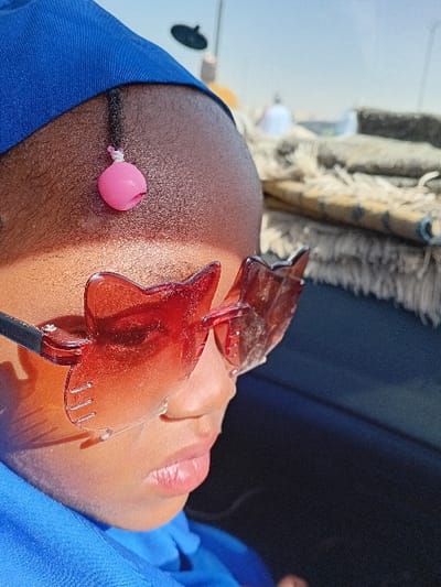An upward-angled close-up depicts a young individual with dark skin, seen in right profile, situated outdoors in Sharada, Nigeria. The subject wears a blue head covering and a blue garment around the neck/shoulder area. Oversized, red-tinted, cat-eye shaped sunglasses with black side frames obscure the eyes. A single, thin braided section of hair on the forehead is adorned with a small, spherical, pink plastic bead secured by a white string. Direct sunlight illuminates the left side of the face, creating highlights on the forehead, nose, and lips. The lips are closed. The blurred background features textured, rolled objects (possibly mats or fabric) with a fringed edge on the right, and a bright, clear blue sky with indistinct dark and light shapes further in the distance.