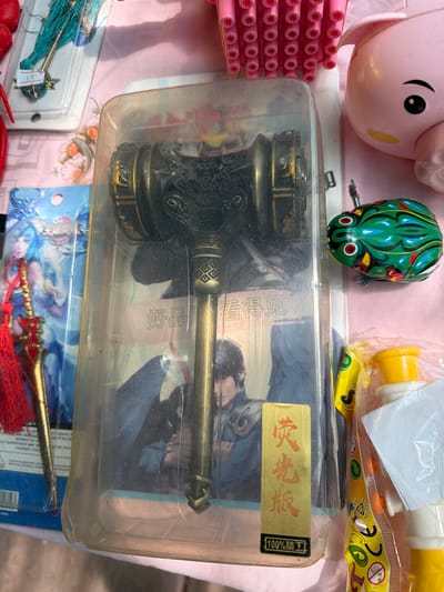 This close-up, overhead image displays a variety of novelty items and toys, likely for sale at a market stall or small shop in Jiejinkou, China. The central subject is a large, ornate, dark-colored (possibly bronze or brass) hammer or gavel-like object, elaborately decorated, enclosed in a clear plastic clamshell case. A gold sticker on this case features Chinese text "荧光版" (Fluorescent version) and "100%精工" (100% finely crafted), with additional text "好品看得见" (Good products are visible) partially seen underneath.

Surrounding this central item, other goods are visible. To the top left, a pink plastic piggy bank or cartoon character head is next to a stack of pink, cylindrical objects. A green wind-up toy frog with red eyes sits at the top center. To the top right, a yellow plastic toy is sealed in a clear bag with visible text including "TOY", "CE", "RECYCLE", and "0-3". At the bottom, two more metallic, decorative items with tassels (one blue, one red) are in clear packaging. The red-tasseled item's packaging includes partial English text such as "NOT SUITABLE FOR CHIL" and "BECAUSE OF SMALL PART", along with "CHINA". The items are laid out on a light-colored fabric, possibly patterned, and the plastic packaging shows some dust or wear. No people are visible in the frame.