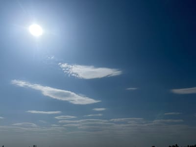 The image displays a clear blue sky with scattered white clouds, illuminated by a bright sun in the upper left corner. The sun is a prominent, blown-out white circle with visible rays extending outwards. Below the sun, a few wispy clouds are present. Further down, a layer of more elongated, flatter clouds stretches across the horizon. The lower part of the image shows a hazy silhouette of trees, suggesting a distant treeline or a landscape feature. There are no people or specific objects identifiable in the scene, other than the natural elements of sky and clouds. The overall impression is of a bright, sunny day in an open environment. The location is specified as Kasri Arifan, Uzbekistan, indicating an outdoor scene in that region.