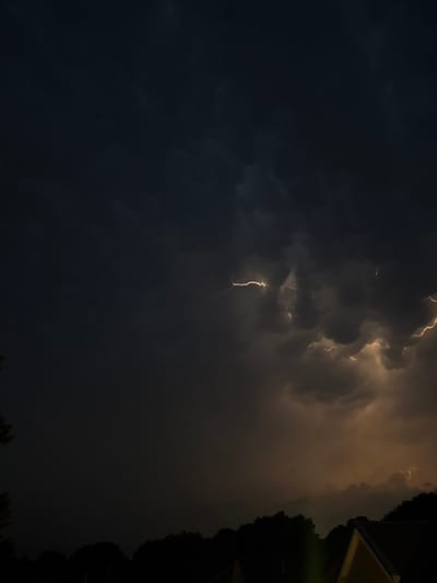 The image depicts a dark, stormy sky with visible lightning strikes illuminating thick clouds. The lower portion of the image shows the silhouetted treeline of a residential area, with the pointed roof of a house in the foreground on the right. There are no people visible in the image. The scene appears to be set at night during a thunderstorm. The location of Putten, Netherlands, is not discernible from the visual information presented in the image.