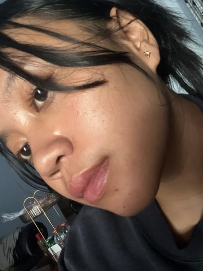 A close-up, angled shot features a young woman's face, capturing her left eye, nose, lips, and ear. Her dark hair is partially obscuring her face, with strands falling across her forehead. She is wearing a dark-colored sweatshirt and a small, gold stud earring in her left ear. The background is dimly lit and out of focus, showing what appears to be a shelf with various items, including bottles and plastic packaging. The lighting suggests an indoor setting. The image does not provide any information about the city of Jambearum, Indonesia, or any context related to that location.