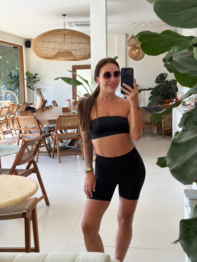 A woman in black exercise attire (sports bra and biker shorts) is taking a selfie in a well-lit, stylish room. She wears sunglasses and a watch, showcasing tattoos on her arm and torso. The room is decorated with large leafy plants, rattan furniture, and woven light fixtures. Other people are seated at a table in the background. The image has been rotated 90 degrees.