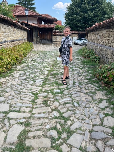 A young boy stands on a cobblestone street in the historic town of Zheravna, Bulgaria. The boy is wearing a black and white patterned t-shirt and shorts, and black sandals. He has a black bag slung over his shoulder and is holding a white piece of paper in his left hand. The street is lined with stone walls and traditional Bulgarian houses with tiled roofs. A silver car is parked in the background. The sky is blue with some clouds. The scene appears to be from a tourist area in Zheravna, known for its well-preserved architecture.