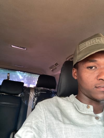 This image depicts a dark-skinned male individual seated within a vehicle, located in Yan Dala, Nigeria. The subject is visible from the mid-chest upwards, occupying the right side of the frame. He wears a light-colored, possibly off-white or light grey, long-sleeved shirt featuring two visible brown buttons on the placket. An olive green baseball cap, adorned with a distressed, flag-like emblem on its front, covers his head. His right eye is open, and he maintains a neutral expression, looking towards the camera. The vehicle's interior shows a light-colored headliner with two oblong overhead lights and a single air vent towards the rear. Directly behind the subject are two black headrests of the second-row seats. The seatback immediately behind the subject is partially covered with clear plastic wrapping. Through the vehicle's rear window, blurred green foliage and bright daylight are visible.