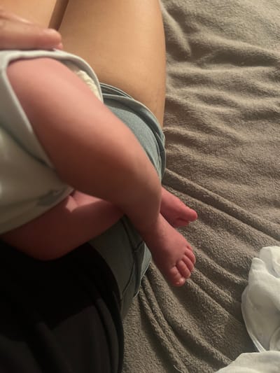 The image shows a close-up view of a person's legs and a baby's feet. The baby's feet are pink and are crossed over each other. The person is wearing denim shorts and a black garment, possibly pants or a shirt. A soft, textured, beige blanket is visible in the background, with some white fabric also present. The lighting suggests it might be indoors, possibly at night or in a dimly lit room, with the light source coming from above and to the left. The overall impression is one of intimacy and tenderness, with the focus on the baby's delicate feet. There is no visible text in the image. The location context of Gorodische, Russia, is provided but not discernible from the visual elements of the image itself.