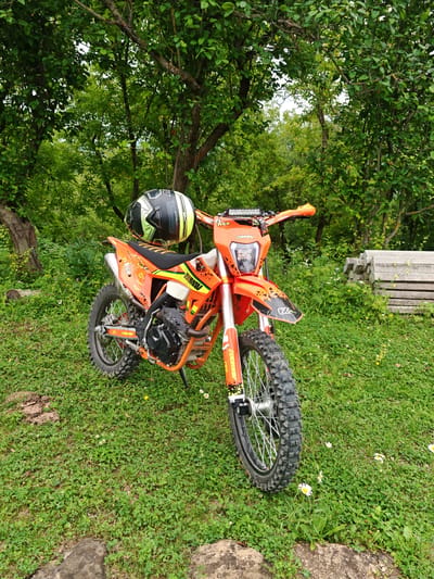 An orange dirt bike is parked on a grassy knoll in a wooded area. A black and yellow helmet rests on the handlebars. In the background, a stack of concrete slabs is visible. The scene is set in the city of Sherdy-Mokhk, Russia.