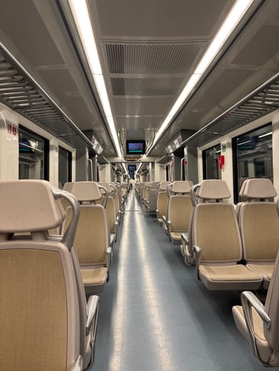 The image shows the interior of a train car, likely a commuter or subway train. Rows of empty seats upholstered in a light brown or beige fabric are visible on either side of a central aisle, receding into the distance. The seats have light gray or beige frames and armrests. The floor is a light blue-gray. The walls of the train car are light gray and feature perforated panels and horizontal lighting fixtures. Windows are present above the seats, with safety and information signage visible near them. Several video screens are embedded in the upper portion of the train car, displaying information. The composition is taken from a perspective at one end of the train car, looking down the length of the aisle. It is not possible to determine whether this is the city of La Marquesa, Mexico based on the provided image.