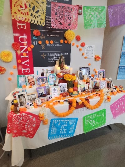 This image depicts a Día de los Muertos *ofrenda* (altar) set up indoors within a building at Seattle University in Seattle, Washington, United States.

The central focus is a rectangular table draped with a white tablecloth, extensively decorated with orange marigold garlands and colorful *papel picado* banners. Numerous framed photographs of individuals (including adults and children) and a pet cat are displayed. The altar features several lit white candles, multiple decorated sugar skulls, a small statue resembling the Virgin Mary or a saint, fresh fruit (oranges, apples), bottles (some appearing as beverages, one as honey), and decorative pompoms.

The background wall is light-colored and features a large black directional sign with white text and icons, indicating locations such as "201 Meeting Room," "Cherry Street Market," and pathways to "Central Campus Upper Mall," "Library Terrace," "Learning Commons," and "The Quad." A vertical red banner displaying "UNIVE" in white lettering hangs on the left. Additional *papel picado* banners in yellow, pink, green, and purple are strung high on the wall, along with numerous small orange and yellow decorative pompoms. A red fire alarm or call box is visible on the wall. No individuals are present in the scene. The floor is a dark, even surface.
