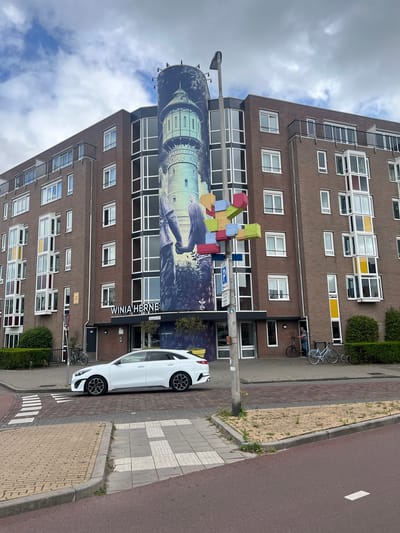 An exterior, medium-shot, eye-level, daylight, full-color, vertical photograph depicts a large, multi-story, brick apartment building with a mural of a water tower and two figures holding hands wrapping around a cylindrical section of the building. The building is located at Winia Herne in Leeuwarden, Netherlands. A white Kia Ceed station wagon is parked at a crosswalk in the foreground. Yellow brick pavers are visible on the sidewalk and a red asphalt street with white lane markings is in the foreground. A streetlamp with multi-colored boxes attached is on the left side of the image. A person is visible standing near the entrance of the building, and two bicycles are parked to the right of the entrance. The sky is partly cloudy.