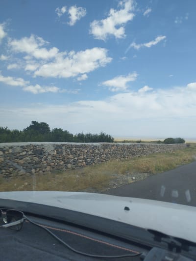 The image, taken from inside a vehicle, shows a long stone wall bordering a road. The wall is constructed of various sized stones and appears to be quite old. Beyond the wall, there is dry grass and sparse vegetation. Further in the distance, there is a line of trees, and beyond that, a hazy horizon that suggests a large open area or body of water. The sky above is blue with scattered white clouds. The foreground of the image shows the dashboard of the vehicle, with a pair of sunglasses and a cable resting on it. The scene appears to be set in a rural or semi-arid environment. The location is identified as O'xum QFY, Uzbekistan.