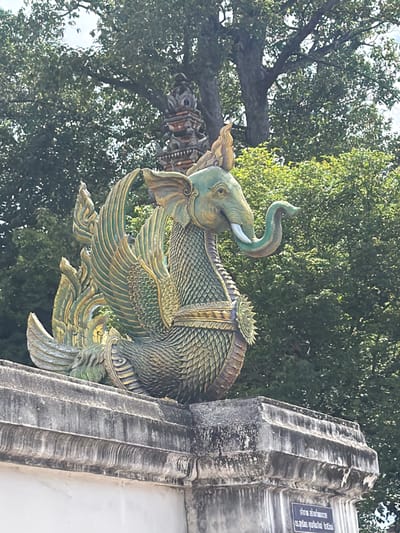 This media file features a decorative sculpture of a mythical creature, possibly a Garuda or a dragon-like being with an elephant's head, adorned with scales and feathered wings. The sculpture is predominantly green and gold, with intricate detailing on its body and wings. It appears to be mounted on a stone structure, which has a weathered, possibly ancient, appearance and is partially obscured by foliage. The background is dominated by lush green trees, suggesting an outdoor setting with ample natural light, likely indicating daytime.

The sculpture itself is the primary subject, posed in a dynamic, almost flying or perching, stance. The elephant's head has a curled trunk, tusks, and large ears, while its body transitions into a serpentine or avian form with prominent, layered wings. The scales on its body are meticulously rendered, and decorative elements, such as a circular crest on its back, add to its ornate nature. The stone pillar on the right side of the frame is textured and has a fluted design. A small sign with text is visible on this pillar, though the text is in a language that is not immediately identifiable but appears to be Thai script, given the location context of Ban Ko Klang, Thailand.

The overall impression is one of a culturally significant artistic piece, likely part of a temple or historical site in Thailand, given the artistic style and location context. The vibrant colors of the sculpture contrast with the muted tones of the stone and the natural greens of the surrounding trees. The weather appears to be clear, allowing for bright lighting. There are no people visible in the frame. The composition focuses on the detailed craftsmanship of the sculpture and its integration with the natural environment and architectural elements.