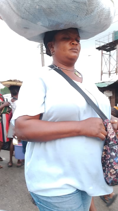 The image shows a medium shot of a Black woman carrying a large sack on her head, walking on a paved street in Agbarho, Nigeria. She is wearing a light blue t-shirt, blue jeans, and a black strap across her chest, which holds a floral patterned bag. She has a silver hoop earring in her left ear and a necklace. In the background, other people and market stalls are visible. The environment appears to be an outdoor market or street scene with a bright, overcast sky.