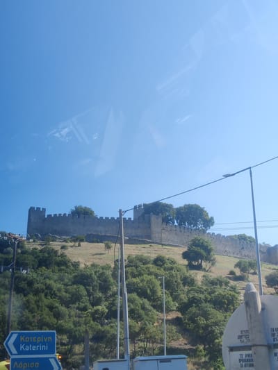 A bright, sunny day with a clear blue sky is captured in this image. The foreground features a road with a blue directional sign pointing left, indicating "Katerini". Below this, another blue sign reads "Larisa". The middle ground is dominated by a large, ancient stone castle wall, appearing weathered and worn, stretching across a grassy hillside dotted with trees. The castle has crenellated battlements and a towering section on the left. A light pole with wires is visible on the right side of the image, in front of the castle. The background is a vast expanse of clear blue sky. The image appears to be taken from a moving vehicle, as evidenced by the reflections on the sky which suggest a window. The castle is identified as being in Neos Panteleimonas, Greece, suggesting it is likely Platamon Castle.
