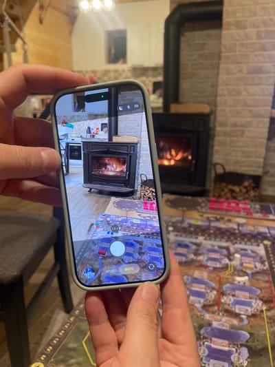 The scene is an indoor environment in Palež, Montenegro. In the foreground, two human hands hold a light-colored smartphone, encased in a pale green protective cover, vertically. The phone's screen displays a live camera feed showing a cast-iron wood-burning stove with a visible fire and a surrounding brick-patterned wall. To the left of the stove on the screen, a modern kitchen area with white cabinets and a stainless steel appliance is visible. On the bottom part of the phone screen, camera interface elements include a white circular shutter button, text labels "ВИДЕО" (Video) and "ФОТО" (Photo) in Cyrillic, and numerical zoom options (0.5x, 1x, 1.8x). An augmented reality (AR) overlay or digital representation of a board game, featuring purple and red structures on a map-like grid, is visible over the floor on the phone screen.
In the background, outside the phone's view, another black cast-iron wood-burning stove with a burning fire is present against a similar brick-patterned wall. To the right of this background stove, a black metal basket contains a stack of cut firewood. A large board game, identical in style to the overlay seen on the phone, is spread out on a dark surface in the lower right, featuring numerous purple, pink, and red game pieces. The room features a light-colored wooden ceiling with exposed beams and a modern light fixture with three spherical bulbs. A dark brown chair or stool is partially visible in the lower left.