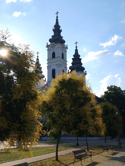 The image depicts the Church of Saint Nicholas in Bač, Serbia, on a sunny day with a bright blue sky and scattered clouds. The church features a prominent white bell tower with a dark spire and a cross at its apex. Two smaller, similarly styled spires flank the main tower, also topped with crosses. The building exhibits arched windows and clock faces on the bell tower. In the foreground, mature trees with yellowing foliage partially obscure the view of the church. A paved walkway runs through a park-like area with benches, and the sun casts long shadows from the trees. The overall scene conveys a peaceful, autumnal atmosphere.