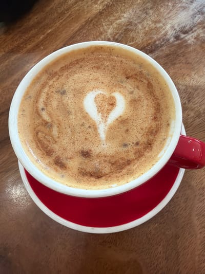 The image shows a close-up, overhead view of a white mug filled with a latte, sitting on a red saucer. The latte has a heart shape created with foam art and is sprinkled with cinnamon. The mug and saucer are resting on a wooden surface, likely a table. The background is a warm, textured wood grain. The lighting suggests it is daytime, possibly indoors in a cafe or restaurant. The overall impression is one of a cozy and pleasant beverage break. There is no visible text in the image. The location context provided is Dallas, United States of America.