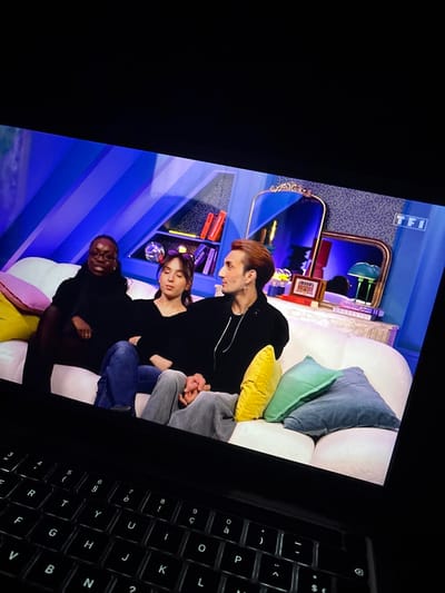 The image displays a laptop screen projecting a television program on the French channel TF1, viewed in a darkened room, from Maisons-Alfort, France. The laptop's backlit keyboard occupies the lower portion of the frame, showing a French-layout QWERTY design.
The program features three individuals seated on a white, rounded sofa in a brightly lit studio setting. From left to right:
1. An individual with dark skin and braided hair, wearing a dark long-sleeved top, looks towards the viewer with a slight smile. A pink cushion is visible to their left.
2. A person with light skin, dark hair, and a purple headband, dressed in a dark long-sleeved top and blue jeans, looks towards the individual on their right. Their left hand is interlocked with the right hand of the third person.
3. An individual with light brown hair, wearing a dark hooded top, grey pants, an earring, and a necklace, looks towards the person on their left. A yellow cushion is positioned between this person and the middle individual.
The studio background is characterized by vertical dark and light blue striped panels, sections with a leopard-print-like texture, and decorative elements. To the left, a shelf displays books with red spines and a disco ball. To the right, a console table holds two ornate gold-framed mirrors, a dark decorative object, and a small green mushroom-shaped lamp. The "TFI" logo is visible in the upper right corner of the screen content.