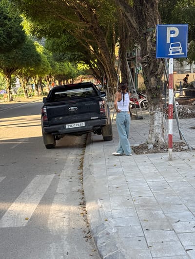 A long shot captures a black Ford Ranger truck parked in Lao Cai Ward, Vietnam. The vehicle, bearing license plate 29K-179.83, is situated on the side of a road lined with lush green trees. A woman dressed in a white tank top and light blue jeans stands near the truck. Adjacent to the truck and the woman, a sidewalk paved with gray stones is visible. A blue parking sign with a white "P" and a bus symbol stands nearby, mounted on a metal frame with red and white stripes.