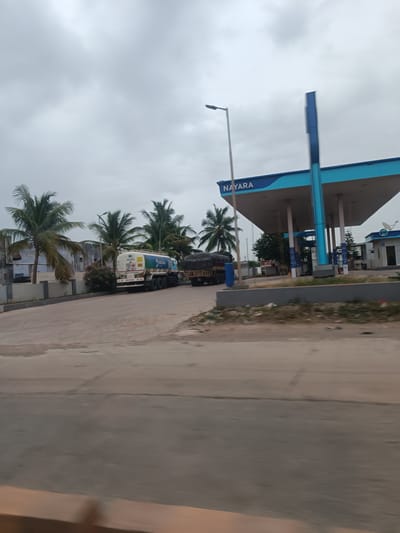 The image captures a Nayara gas station in Bethamcherla, India, under an overcast sky. Two large trucks are visible, one a white tanker truck with "NAYARA" branding and hazard symbols, and another truck loaded with dark materials and covered with a tarp. Palm trees are scattered throughout the scene, and a lamppost stands to the left of the gas station's canopy, which is also branded with "NAYARA." The gas station features prominent blue and white structures. The foreground shows a paved road with some debris and puddles, suggesting recent rain or a generally damp environment. The overall impression is one of a typical day at a roadside establishment in India.