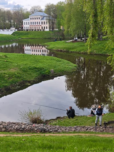 The image depicts a waterfront scene in Uglich, Russia. In the foreground, two people are positioned near the water's edge, along a bank reinforced with rocks. One individual kneels, holding a fishing rod, while the other stands. Further back along the waterfront, people are visible gathered on a grassy area, and others appear on a platform or deck extending out toward the water. Across the water, a pale-colored building with a gray roof can be seen. The building is mirrored in the water. Trees are interspersed throughout the scene, and the sky is overcast.