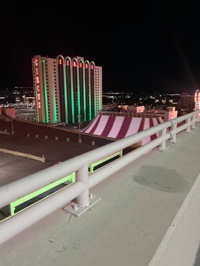 The image depicts a nighttime aerial view of the Circus Circus Hotel and Casino in Reno, Nevada, United States. The vantage point is from a concrete rooftop, likely a parking garage, featuring a white metal guardrail in the immediate foreground. The concrete surface exhibits fine cracks and a discolored patch.

In the midground, a large tent structure with a sloped roof displaying alternating white and magenta/purple stripes is visible. Adjacent to this tent is an open-air concrete area, likely another level of a parking structure, marked with distinct dark tire skid patterns, including circular "doughnut" marks. Several concrete bollards are spaced across this area. A smaller building with flagpoles stands near the tent.

The background is dominated by the multi-story Circus Circus hotel tower, illuminated by vertical green neon lights along its facade. Red neon letters spelling "CIRCUS" are prominently displayed both vertically on one side of the tower and horizontally across its top, with each letter at the top framed by an arched architectural element. Beyond the hotel, the illuminated cityscape of Reno extends into the dark night, with distant lights marking buildings and a faint outline of hills or mountains against the horizon. A light beam emanates from the far right of the frame. No individuals are visible in the scene.