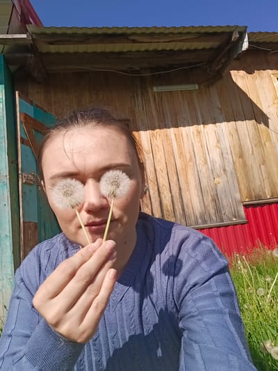 A woman in a blue sweater holds two dandelions over her eyes outdoors in Gabdyukovo, Russia. Behind her, a wooden building with a corrugated metal roof is visible, along with green grass and a clear blue sky. The building has a section painted red, and another section painted teal. The woman is looking directly at the camera.
