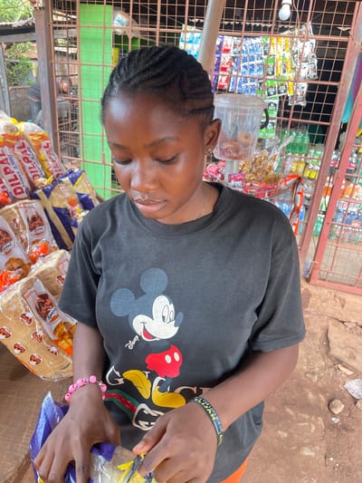 A woman with braided hair wearing a Mickey Mouse t-shirt is at a stand in Otukpa, Nigeria. She has a pink beaded bracelet on her right wrist and a black and yellow wristband on her left that reads “Elijah is the Lord God of”. She is bagging bread with labels “Special” and “Dilpa Family Jumbo” on a wooden table. Behind her is a metal grid displaying various items.