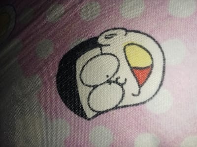 This image depicts a fragment of a patterned textile object located within the Umsobomvu Local Municipality, South Africa. The object is a light pink fabric adorned with white polka dots, displaying a stylized, two-dimensional cartoon character. This character, resembling a penguin, features a white body with a black outline and a solid black section on its left. It has two prominent white oval eyes, each with a small black pupil, and a subtle black curved line below them. An open mouth or beak is visible on the right side of the character's head, colored yellow on the upper portion and red on the lower, both outlined in black. The fabric's weave is discernible, and the scene is illuminated by uneven ambient light, creating a slight gradient from darker on the left to lighter on the right. There are no individuals, actions, or dynamic interactions captured.
