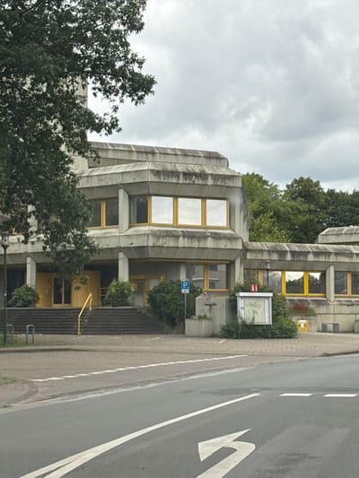 The image shows a modern, Brutalist-style building with a concrete facade and yellow-framed windows. The building has multiple levels and angular architectural features. A wide set of stairs with a yellow railing leads up to the entrance on the ground floor, which appears to be yellow. To the left of the stairs, a large tree with dark green foliage partially obscures the building. In front of the building, a paved area with some green shrubbery and a notice board is visible. A blue parking sign is also present. The building is situated in front of a line of lush green trees. A street with asphalt and white markings is in the foreground, suggesting an urban or suburban setting. The sky is overcast with gray clouds. This building is the Bürgerzentrum (Civic Center) in Gronau, Germany.
