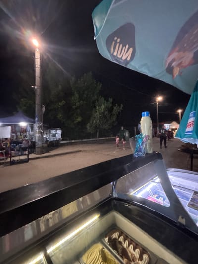 The image depicts an outdoor, nighttime scene in the city of Kostievo, Bulgaria, likely a street market or fair. In the immediate foreground, a black-framed ice cream display freezer is visible, with its glass lid open. Inside, multiple containers of ice cream are present, including one with chocolate and white cream swirls, and another with a yellow, solid appearance. The interior of the freezer is illuminated by bright linear LED lights.
Resting on the black frame of the ice cream display, to the right, are a tall, opaque, light-yellow bottle with a blue screw cap, and a clear glass or plastic holder containing multiple colorful plastic spoons or stirrers.
Above and to the right, a large portion of a light-blue or teal market umbrella is visible, featuring white scalloped edges. On its surface, a dark logo with the inverted text "iiuv" (likely "nuun" when viewed correctly) is present. Another partially visible logo, showing "es", is on the lower right section of the umbrella.
The midground features an unpaved or dirt road/path illuminated by various light sources. A prominent utility pole stands centrally on the left, topped with a bright, glowing street lamp emitting significant light flares. To the left of the pole, a vendor stall is set up under a dark blue tent, displaying various items for sale. To the right of the pole, another smaller display or stall is present.
Further down the path, several individuals are visible. Two smaller figures, likely children, one in a pink dress, are walking away from the camera, near a taller, dark-clothed adult figure. In the background, additional street lamps provide further illumination, and more people are discernible along the path. Dense, dark green trees and foliage line the background, indicating a natural or planted area. The sky is dark, consistent with the nighttime setting.