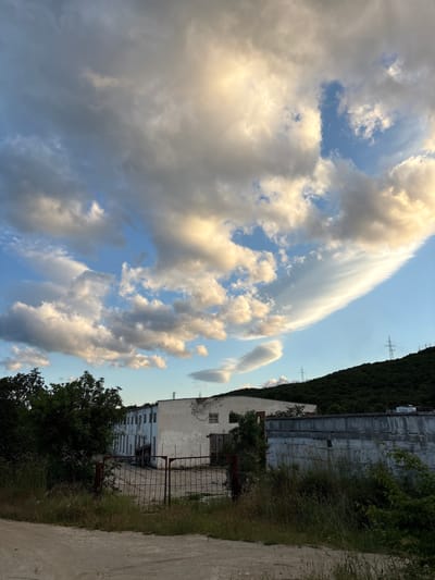 The image depicts an outdoor scene with an industrial building and a lush, tree-covered hillside under a dramatic sky filled with clouds. In the foreground, a dirt path or road leads towards an area with overgrown grass and weeds. A rusty metal gate stands open, revealing a concrete yard in front of a long, white, multi-story building. This building shows signs of disrepair, with peeling paint and some windows boarded up or missing. Adjacent to this building is a lower, concrete structure with a weathered facade.

Behind the buildings, a steep, dark green hillside is densely covered with trees. Several tall electrical pylons are visible on the crest of the hill. The sky above is a striking display of dynamic clouds. Large, fluffy cumulus clouds are illuminated by the setting sun, casting golden and white light, contrasting with areas of blue sky. A distinctive, wispy lenticular cloud stretches horizontally across the upper right portion of the frame. The overall impression is one of an industrial area bordering a natural landscape, captured during the golden hour before sunset.