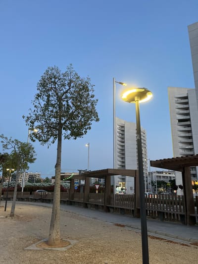 The image captures an urban public space at dusk or early evening, characterized by a clear, deep blue sky. The scene is dominated by a prominent, modern streetlight with a circular LED fixture emitting bright yellow light, positioned centrally. Its tall, slender pole extends downwards, with a smaller, more conventional streetlight fixture visible attached to it and another one further to the left.
To the left, a mature tree with a substantial trunk and a full canopy of green leaves stands, its branches reaching into the sky. Further trees, also with slender trunks, are visible towards the lower right. The ground consists of a gravel or dirt path, edged by concrete, with a round metal grate visible at the base of one tree.
In the mid-ground and background, contemporary multi-story buildings rise against the sky. These structures feature numerous windows and distinctive architectural elements such as slatted or louvered facades, suggesting apartment complexes or office buildings. Some windows in the buildings are illuminated, indicating the time of day. Wooden pergolas or shelters with slatted roofs and benches line the pathway to the right, suggesting a recreational area.
No people are visible, and there is no specific activity taking place, lending a calm and tranquil atmosphere to the scene. The weather appears clear and cloudless.
Notable details include a small, red triangular sign with a white border in the distance, partially obscured by a wooden structure. On the prominent streetlight pole, a white label with some dark text is visible, with faint numbers resembling "1450" and potentially "mm" below it, though the text is largely unreadable.
