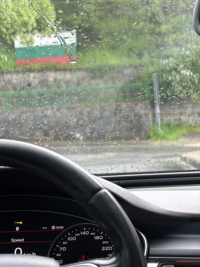 The image is taken from inside a car, likely stationary given the speedometer reads 0 km/h. Raindrops are visible on the windshield, partially obscuring the view of the exterior. The exterior view shows a road or path alongside a green fence-like structure and some greenery. The red and white colored object is possibly a sign. The car's dashboard and steering wheel are prominent in the foreground. Given that this location is in Davidkovo, Bulgaria, there is not enough information to determine the specific spot shown in this photo.