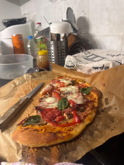 A freshly baked homemade pizza is the central focus of this indoor kitchen scene, presented on crumpled brown parchment paper. The pizza is generously topped with melted mozzarella cheese, sliced red tomatoes, basil leaves, red bell peppers, and what appear to be mushrooms and possibly onions. Its crust is golden brown, indicating it's just out of the oven. A kitchen knife with a wooden handle rests alongside the pizza, suggesting it is about to be cut and served.
To the left of the pizza, a white kitchen towel with blue illustrations and text is visible. Partial text like "opm" and "KAA MIO" can be discerned on the towel. The background reveals a cluttered kitchen environment, typical of a home setting. Various kitchen utensils and containers are visible, including a metal utensil holder, white mugs, a metal strainer, and several plastic bottles containing liquids, possibly cooking oil or water. A clear plastic measuring cup and a larger white plastic bucket are also present. The overall impression is that of a casual home kitchen, likely in the evening given the artificial lighting and the warm glow on the pizza. No people are visible.