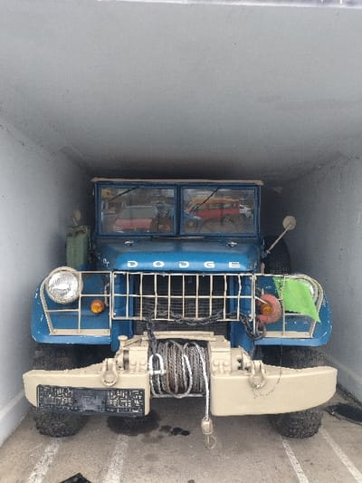 This image depicts the front view of a vintage blue Dodge utility vehicle positioned within an enclosed, narrow, light-colored space in Bitburg, Germany. The vehicle features "DODGE" lettering on its hood, a vertical-bar grille, two circular headlights, and amber turn signals. A tan-colored front bumper assembly is equipped with a central winch containing coiled cable and two visible tow shackles. A black license plate holder is affixed to the left side of the bumper, and a small green flag-like object is attached near the right side of the grille. The vehicle has a split windshield, reflecting external objects including red and yellow/orange vehicles. The floor beneath the vehicle is a light-colored hard surface, exhibiting dark fluid stains near the front bumper. Faint white floor markings are present. The surrounding walls and ceiling of the enclosure are off-white or light grey. No individuals are visible.