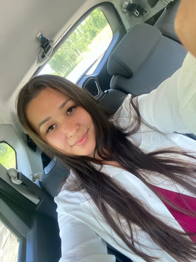 A young woman with long, dark hair and light skin is taking a selfie in the back seat of a car. She is wearing a white shirt over a pink garment. Behind her, car seats and a seatbelt are visible. Outside the window, trees and greenery suggest a rural or suburban setting. The image does not provide enough information to determine if the location is Yuldybayevo, Russia.