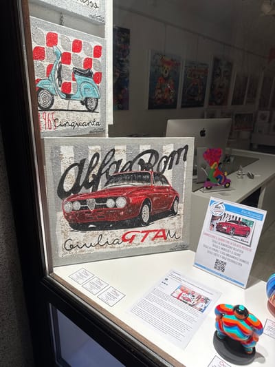 The image shows the interior of a shop window in Orta San Giulio, Italy (latitude: 45.799, longitude: 8.406). The window is displaying a variety of artwork, including a painting of a red Alfa Romeo Giulia GTA, a painting of a blue Vespa scooter, and a colorful ceramic sculpture of a person. The window also has a sign with information about the shop and its services. The shop appears to be an art gallery. There are two small, framed, blank canvases on the bottom of the window along with several brochures and leaflets, one of which displays a photo of three people.  The shop window is in a room with several framed artworks behind it, and a desk with a computer and some papers.  There is no one visible in the image.  The overall mood of the image is bright and welcoming.