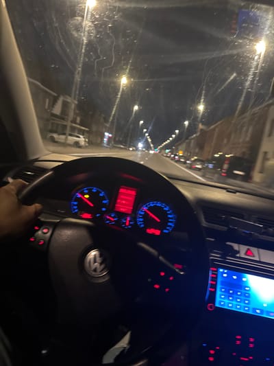 The image shows a first-person perspective from inside a car driving at night in Waregem, Belgium. The dashboard, including the steering wheel and a digital display, is visible. The windshield has streaks of water, indicating rain. Streetlights and buildings line the road ahead. The overall scene depicts nighttime driving through a city.