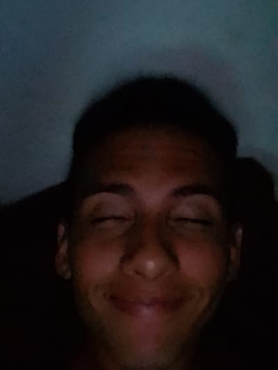 The image features a close-up, dimly lit portrait of a person, likely male, against a dark and muted blue-grey background. The subject is positioned centrally, facing directly towards the camera. He has dark, short hair and appears to be smiling or smirking with his eyes closed. His facial expression conveys a sense of contentment, amusement, or perhaps drowsiness.
Notable details include the low light conditions, suggesting it might be nighttime or a dark indoor setting. The lighting illuminates his face unevenly, highlighting his features such as a prominent nose, full lips, and dark eyebrows, while casting shadows that add depth to the portrait. His skin tone appears warm, and some texture is visible on his face. The background is indistinct, appearing to be a plain wall or soft surface in a dark room. No objects are clearly visible besides the person. The overall impression is an intimate, candid moment. There is no visible text in the image. The location context provided is El Espinal, Venezuela.