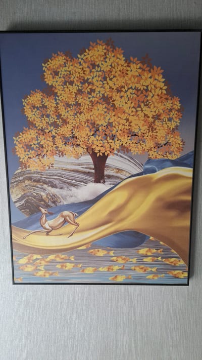 This is a close-up, eye-level, vertical shot of a framed piece of art hanging on a textured, light grey wall. The artwork depicts a stylized scene with a large tree with golden-orange leaves dominating the upper half. A brown tree trunk emerges from white, cloud-like formations and appears to be rooted in swirling, marbled earth tones. Below the tree, a light brown deer with delicate features is positioned on its hind legs, looking upwards. In front of the deer, a large, flowing drape of golden-yellow fabric curves across the landscape. The lower portion of the artwork shows a body of water with numerous small, golden fish swimming. The background of the artwork is a deep blue. The lighting is even, indicating artificial light. The location is not identifiable within the image itself; it is a piece of art displayed on a wall, without any contextual elements indicating a specific city or geographical location.