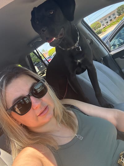In Lake Zurich, United States of America, a woman with blonde hair and sunglasses takes a selfie in the driver's seat of a car, with a large black dog in the back seat. The dog is sitting on the passenger seat, looking towards the camera with its tongue slightly out. The woman is wearing a green tank top and a silver necklace with a pendant. The car's interior, including the grey leather seats and the steering wheel, is visible. Outside the car, through the windows, a suburban environment with buildings, trees, and parked cars can be seen under a clear blue sky.