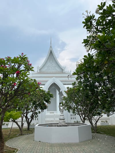 The image shows a low-angle view of a white building with ornate, pointed roofs in Muang Pattaya, Thailand. The architectural style is possibly Buddhist-inspired, given the white color and intricate detailing. A pair of Plumeria trees with both white and reddish-pink flowers frame the view of the building. The ground is paved with greenish-grey bricks. The sky is visible between the buildings and trees, with some cloudy patches. No people are visible.