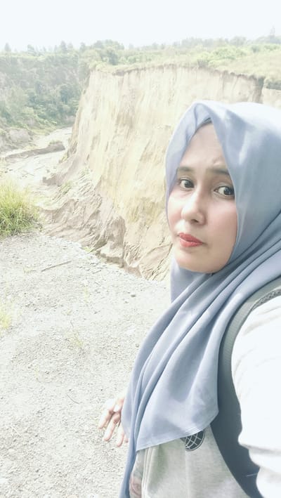 This media file is a selfie taken by a woman wearing a light blue hijab. She is positioned on the right side of the frame, with her face partially visible and looking towards the camera. Her makeup includes red lipstick and subtle eye makeup. She is wearing a grey hoodie underneath the hijab and a backpack is visible on her shoulder. Her left hand is extended, with fingers slightly splayed, resting near the bottom of the frame.

The background of the selfie depicts a natural landscape. Immediately behind the woman is a textured, rocky ground with sparse vegetation. Further in the background, there is a dramatic cliff face with layered soil and sand, indicative of erosion. Green trees and foliage are visible atop the cliff and in the distance, suggesting a somewhat hilly or mountainous terrain. A dry riverbed or gully is visible at the base of the cliff. The lighting suggests it is daytime, possibly late morning or afternoon, with a slightly hazy or overcast sky contributing to the soft, diffused light. The overall setting appears to be a natural, rugged outdoor environment.

The woman's expression is neutral, perhaps slightly thoughtful. The image captures a moment of personal reflection or exploration within this natural setting, likely in Kebunbaru, Indonesia, as indicated by the location context. No visible text is present in the image.